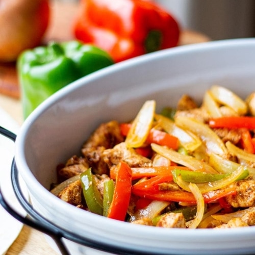 Baked Chicken Fajita mix in white casserole dish on butcherblock countertop with bell peppers and onions in background.