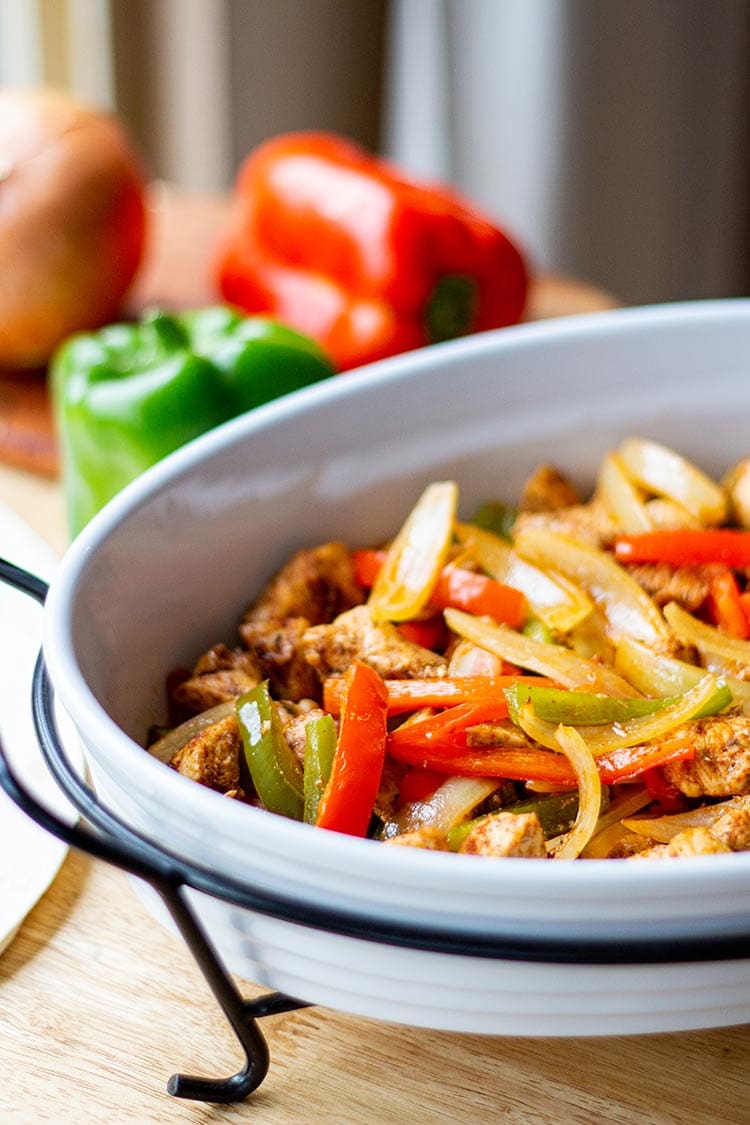 Baked Chicken Fajita mix in white casserole dish on butcherblock countertop with bell peppers and onions in background.