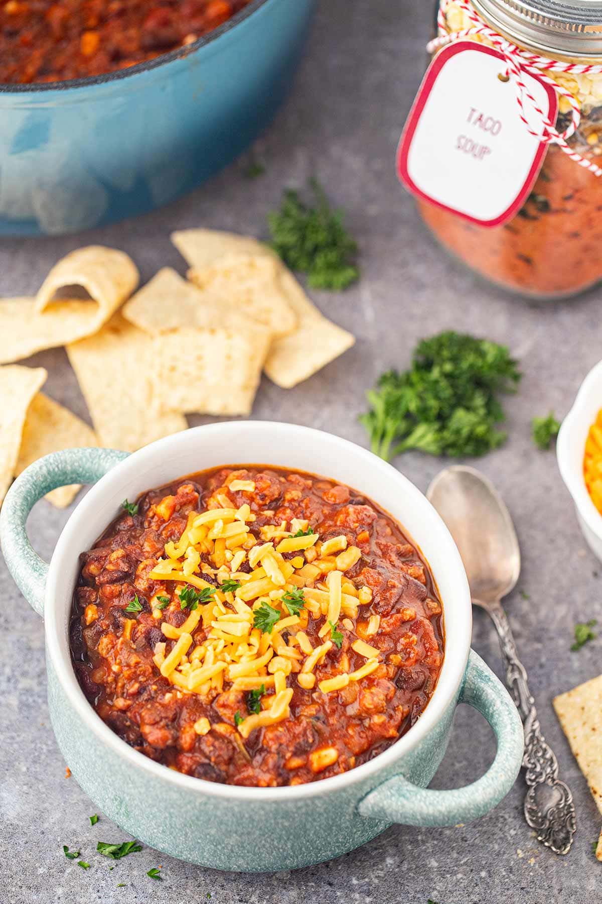 Table scene showing bowl of Taco Soup in a Jar made into soup, surrounded by ingredients, pot of soup, and meal in a jar.