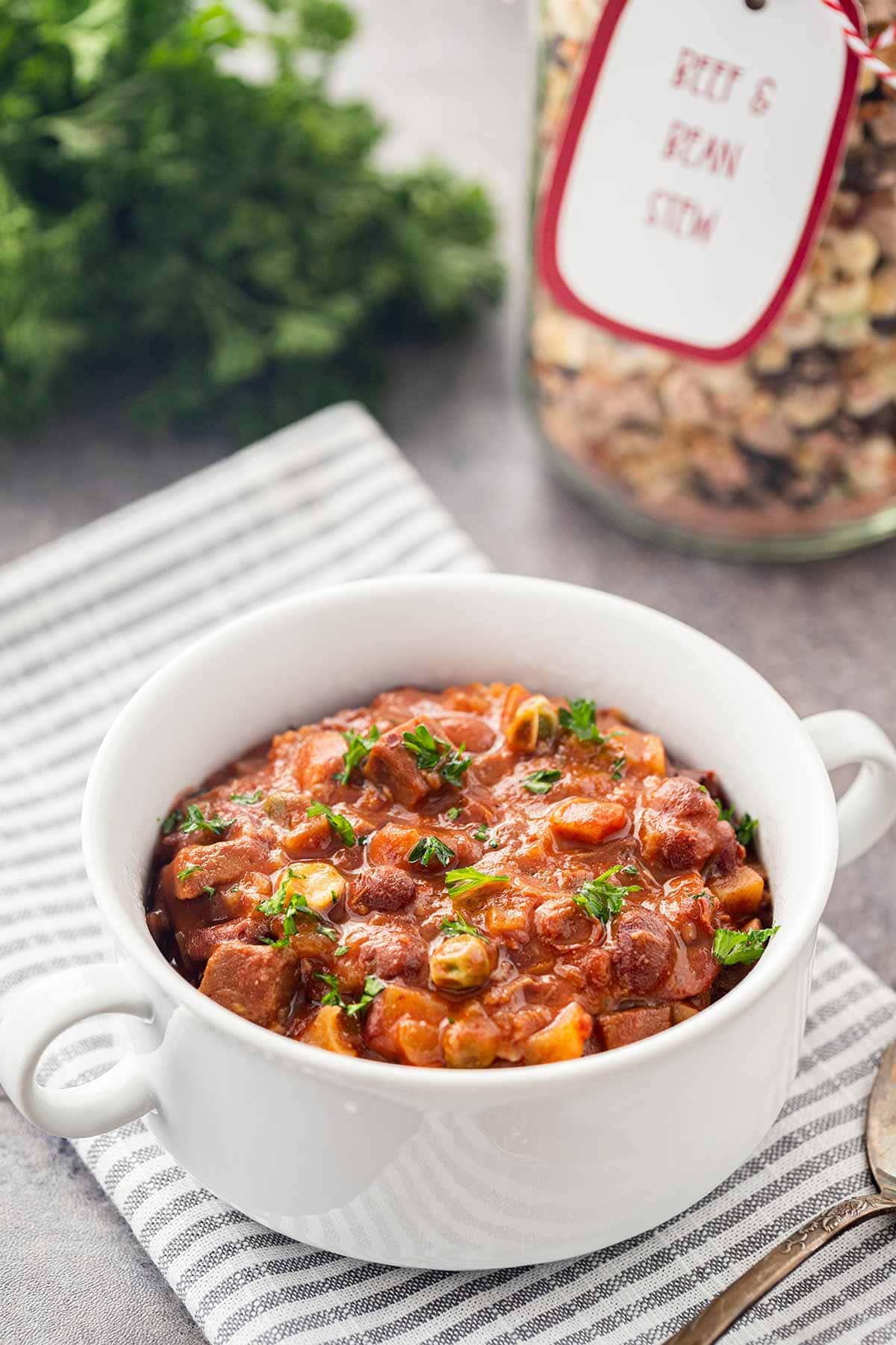 A white bowl of Beef And Bean Stew Meal in a Jar made up and ready to eat, sitting on gray striped napkin on a counter with jar and fresh parsley in background.