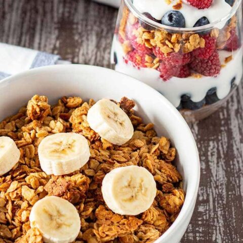 Bowl of Easy Peanut Butter Granola cereal topped with sliced bananas, with a fruit and yogurt granola parfait in the background.
