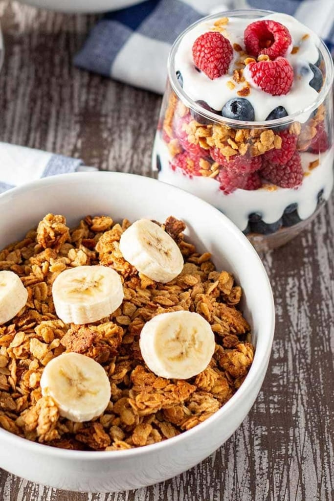 Bowl of Easy Peanut Butter Granola cereal topped with sliced bananas, with a fruit and yogurt granola parfait in the background.