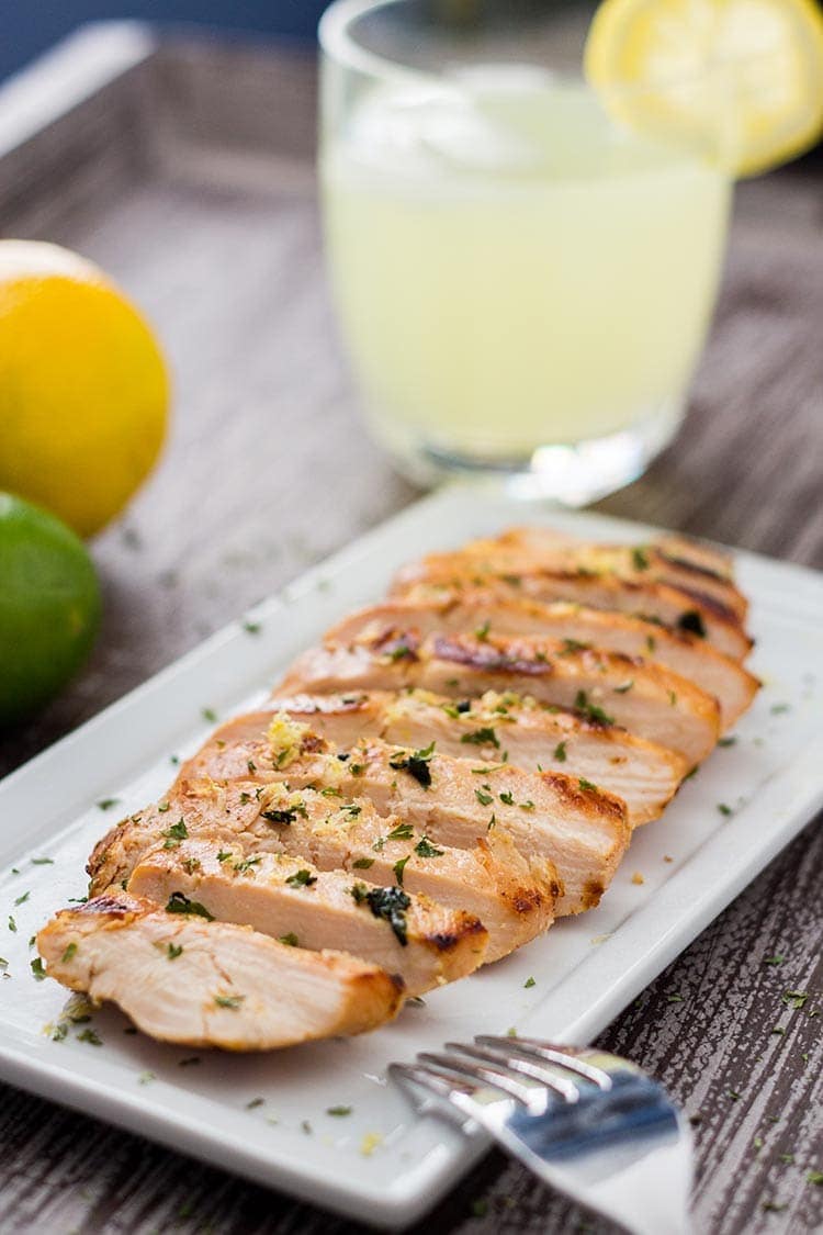 Grilled Lemonade Chicken sliced on a platter and garnished with lemon zest and parsley, with glass of lemonade in background.