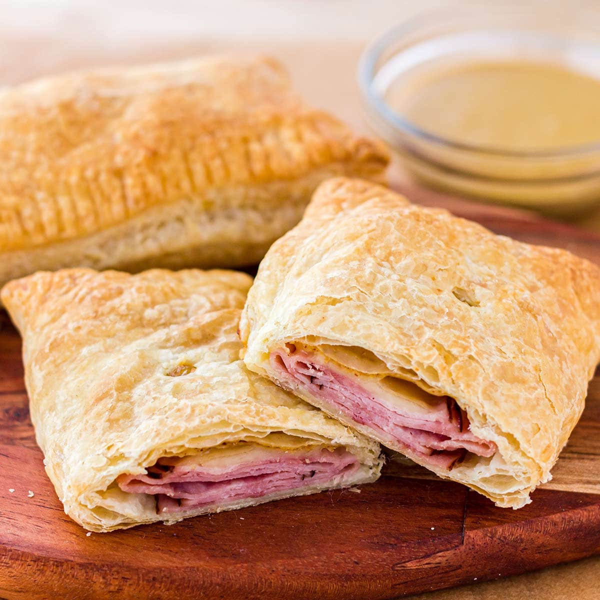 Homemade ham and cheese hot pockets made with puff pastry, on wooden cutting board with mustard dipping sauce in background.