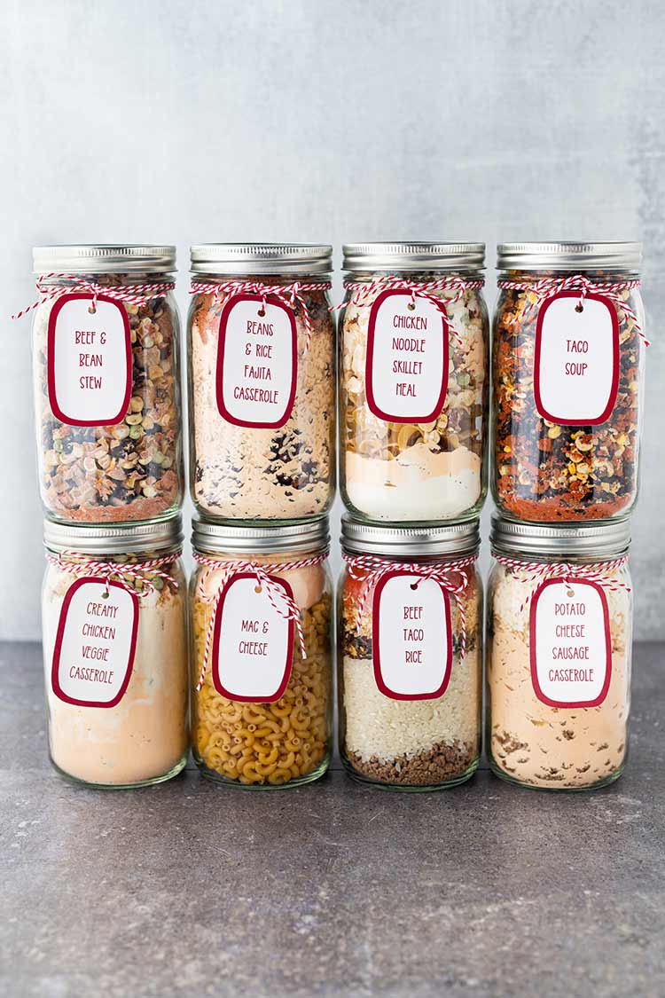Picture of eight meals in a jar stacked on countertop, in mason jars, with labels and gift tags.