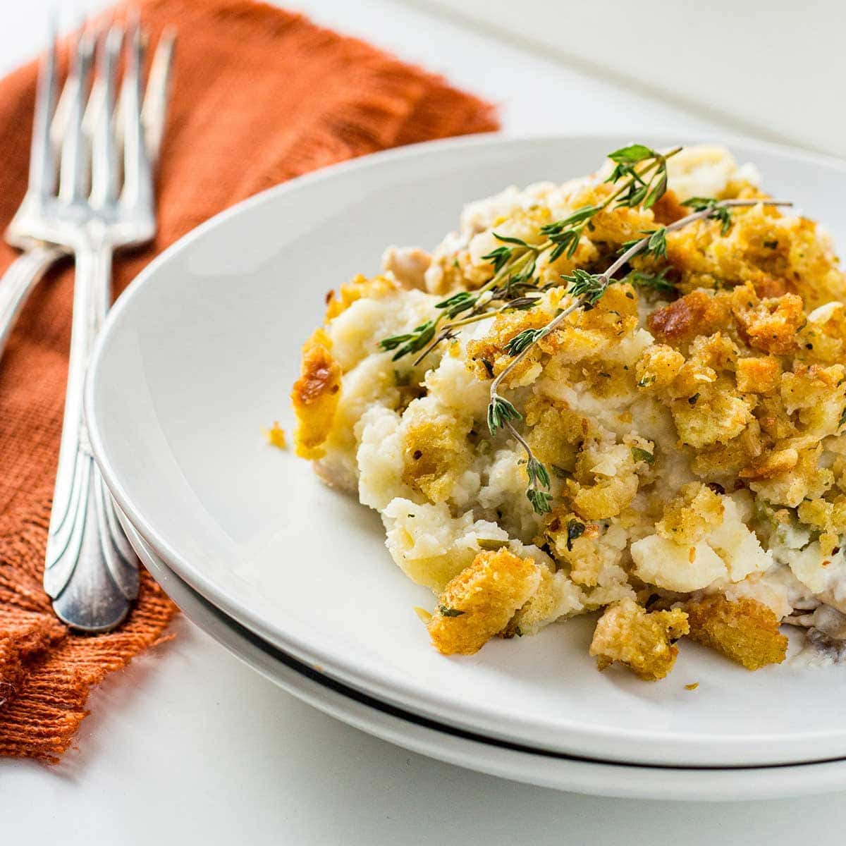 Thanksgiving Casserole dished on a white plate, garnished with sprigs of fresh thyme, with orange napkin and forks in background.