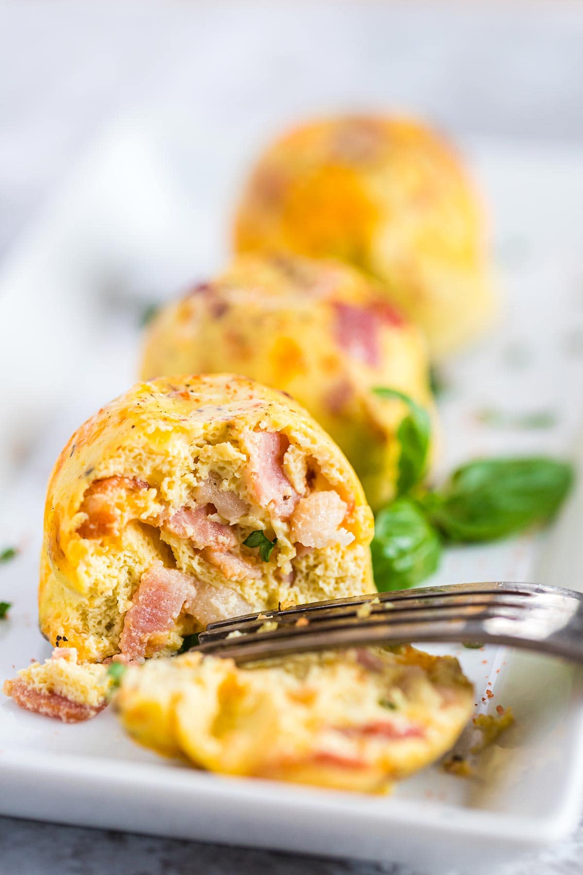 Three bacon cheddar egg bites on white plate with fresh basil and fork.