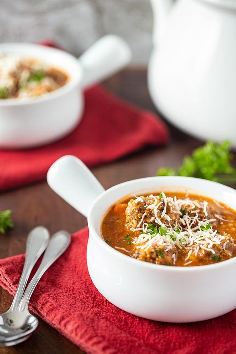 Instant Pot Hearty Meatball Soup in a white soup dish garnished with shredded cheese and fresh parsley in table setting