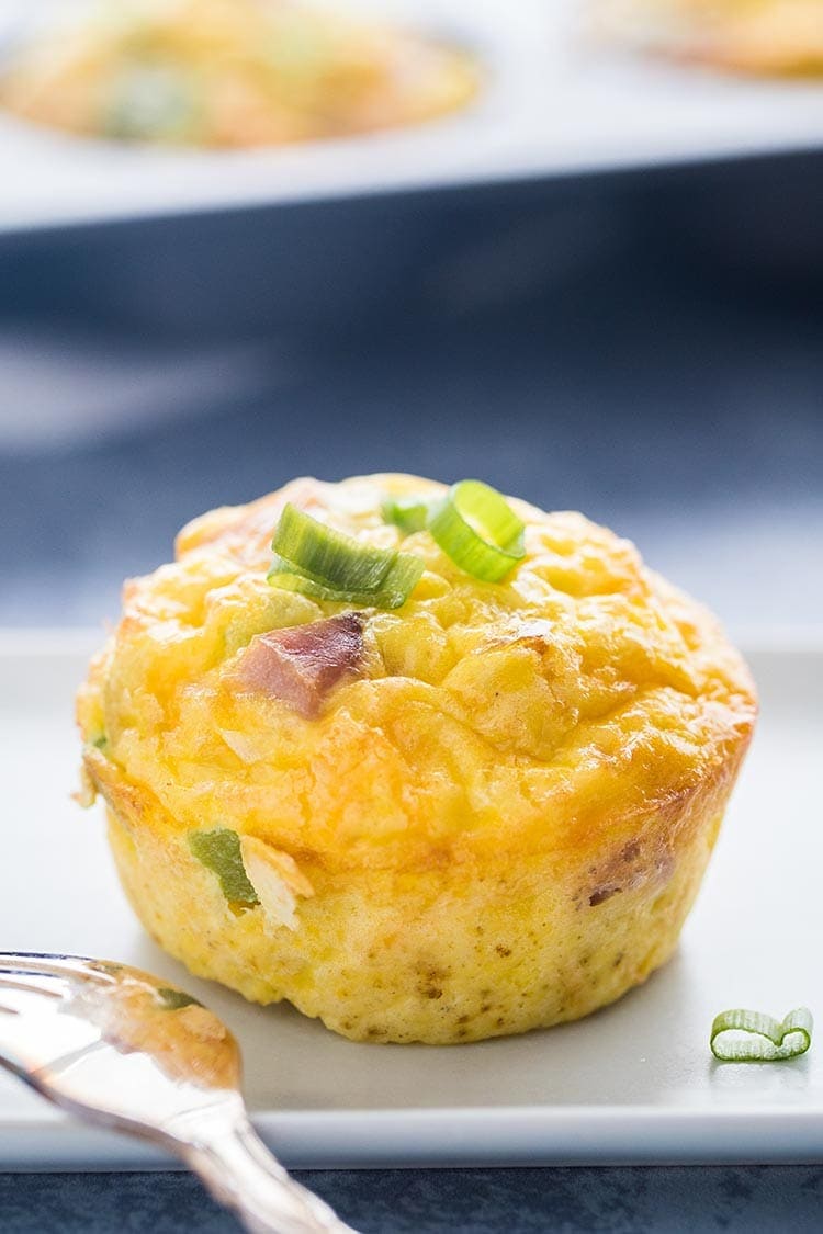 A single Denver Omelet Egg Muffin on a white plate on a blue background in front of a tray of egg muffins.