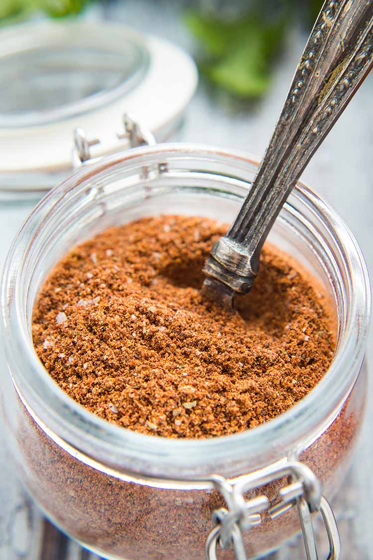 A closeup shot of Taco Seasoning Mix in a small glass jar with fliptop lid and measuring spoon sticking out