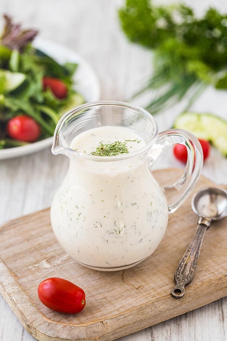 Small glass pitcher of homemade ranch dressing mix sitting on a cutting board with salad and fresh greens in the background.