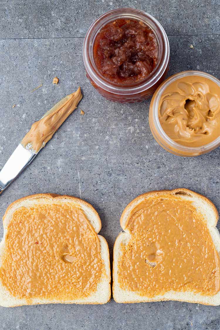 Two pieces of bread on a counter with peanut butter spread on both pieces, with jam and peanut butter in background.