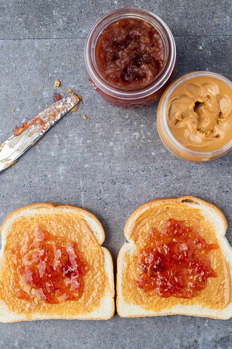 Two pieces of bread on counter, with peanut butter and jam on both pieces.