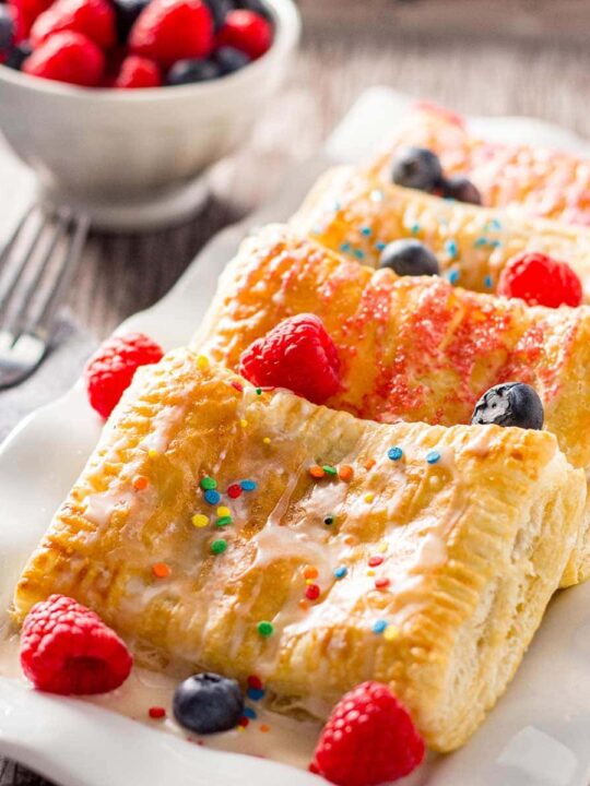 Four baked breakfast pastries layered on a white plate, garnished with fresh fruit, icing, and sprinkles.