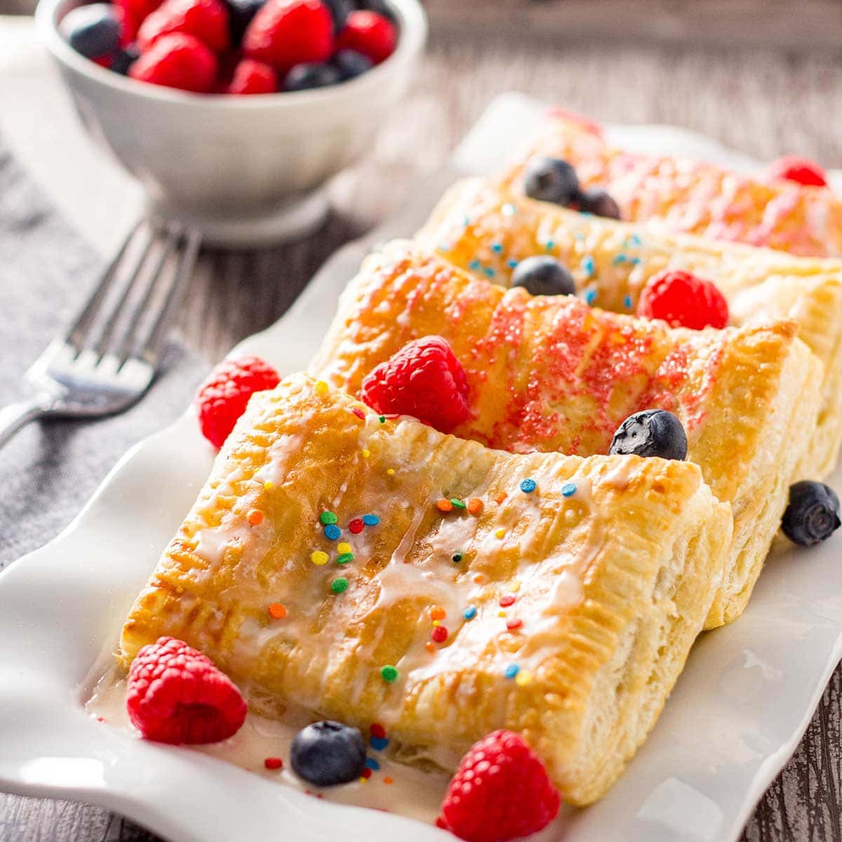 Four baked breakfast pastries layered on a white plate, garnished with fresh fruit, icing, and sprinkles.