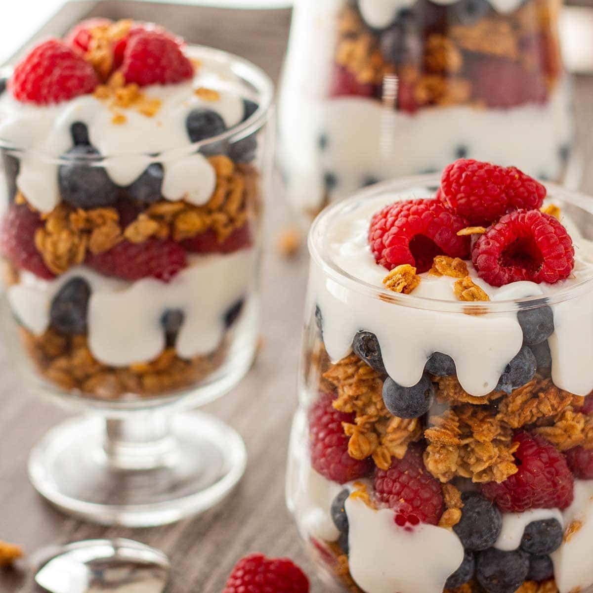 Three glasses, staggered, filled with layers of granola, yogurt, and fresh fruit, garnished with additional fruit and granola, on a gray wooden tray.
