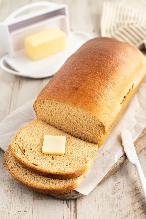 Loaf of honey wheat bread sitting on cutting board, with two slices cut off front with pat of butter.