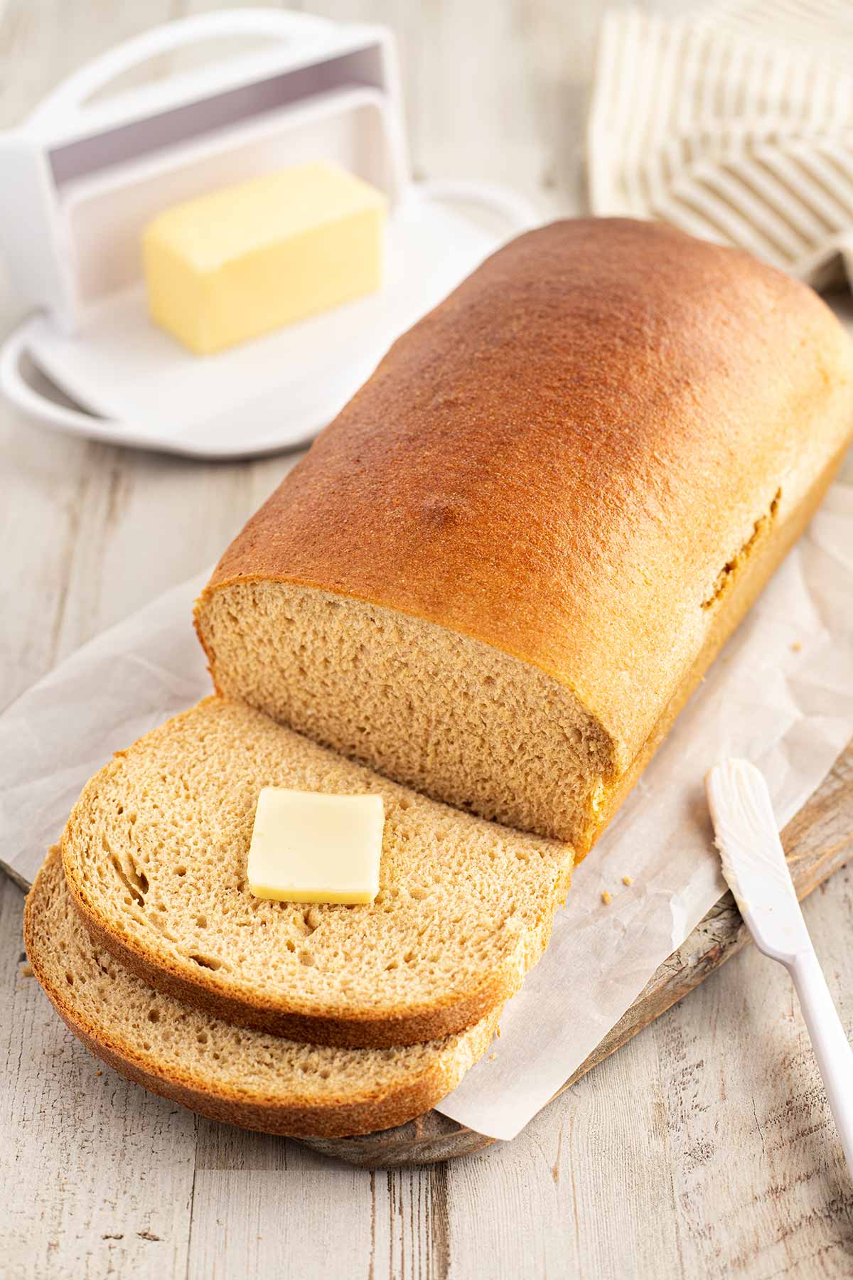 Loaf of honey wheat bread sitting on cutting board, with two slices cut off front with pat of butter.