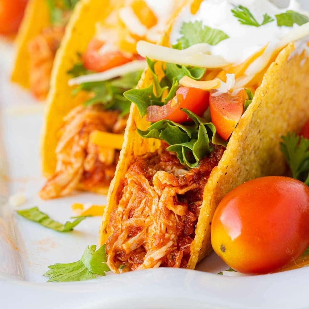 A close up of three tacos made with Instant Pot Salsa Chicken, lettuce, tomatoes, cheese, sour cream, and garnished with cilantro, staggered on a white plate.