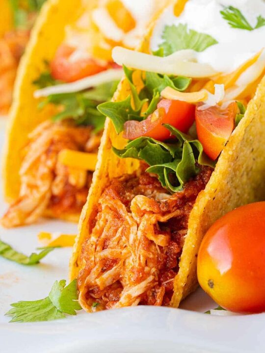 A close up of three tacos made with Instant Pot Salsa Chicken, lettuce, tomatoes, cheese, sour cream, and garnished with cilantro, staggered on a white plate.
