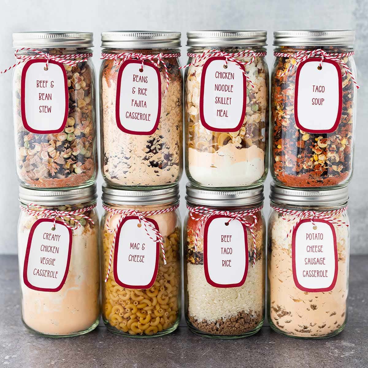 Eight quart jars, stacked on top of each other, in front of gray background, with printed labels tied to the outside of each jar and each filled with the dry ingredients to make a meal in a jar.