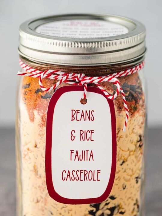 Mason jar, on gray background, filled with the dry ingredients used to make Bean & Rice Fajita Casserole, sealed and a printed label tied to the rim of the jar.