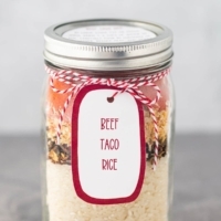 A quart mason jar, on a gray background, filled with the dry ingredients for Beef Taco Rice, with a printed label tied to the top of the jar.