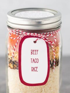 A quart mason jar, on a gray background, filled with the dry ingredients for Beef Taco Rice, with a printed label tied to the top of the jar.