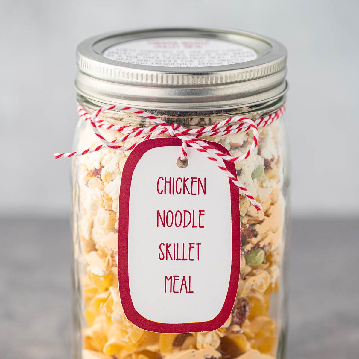 Quart mason jar, on gray background, filled with the dry ingredients used to make Chicken Noodle Skillet Meal, with a printed label tied to the outside of the jar.