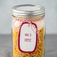 A quart mason jar, on a gray background, filled with the dry ingredients needed to make this Mac & Cheese meal in a jar, with a printed label tied to the outside of the jar.