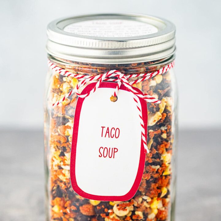 A quart mason jar, on a gray background, filled with the dry ingredients needed to make Taco Soup, with a printed label tied to the outside of the jar.