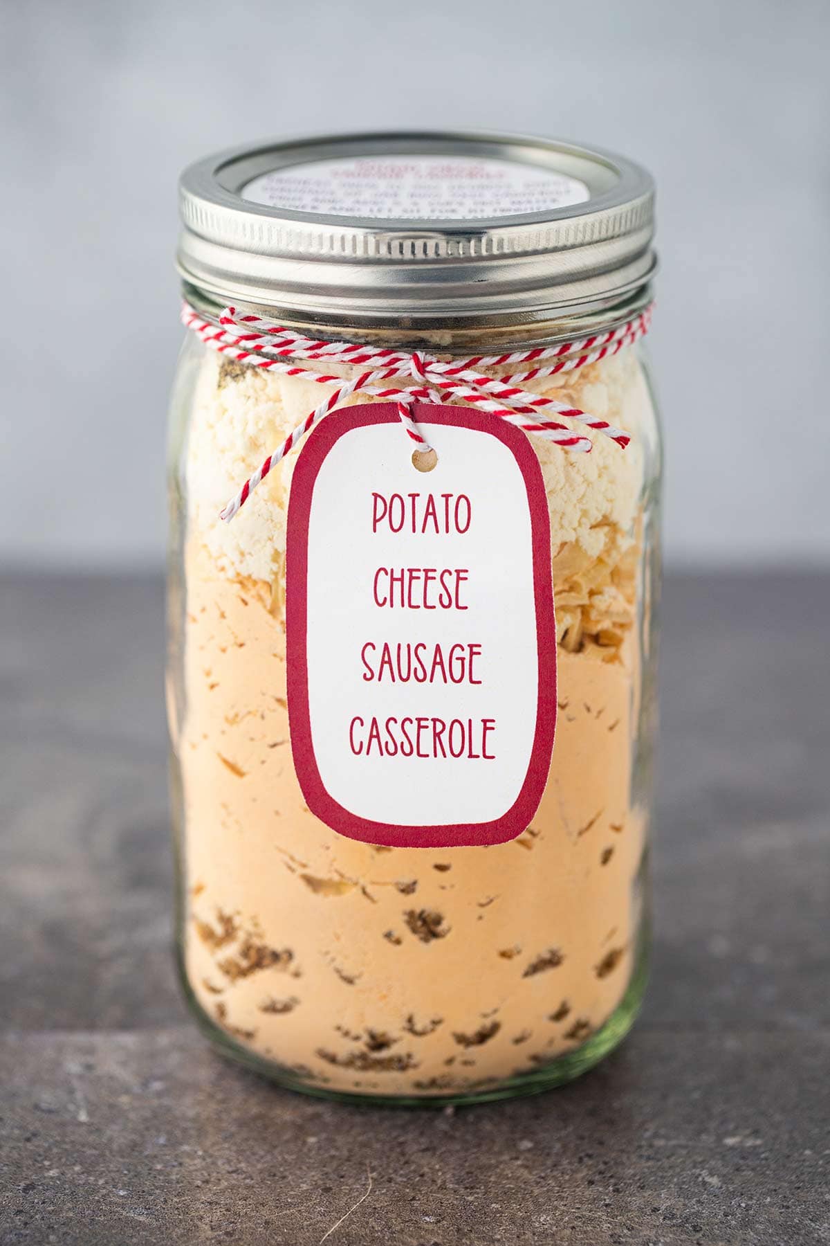 Quart mason jar filled with the dry ingredients to make Potato Cheese Sausage Casserole, sealed and with a printed label tied to the front, with gray background.
