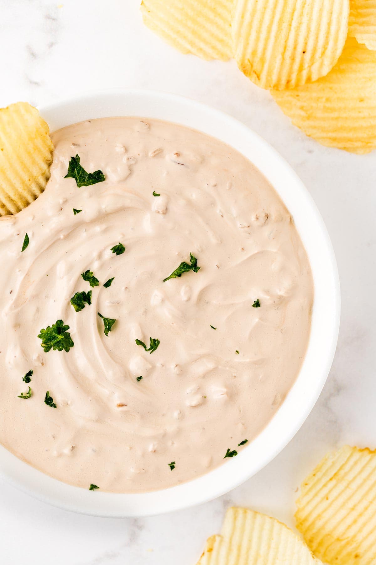 Overhead shot of french onion chip dip in white bowl, surrounded by potato chips and garnished with fresh chopped green parsley.