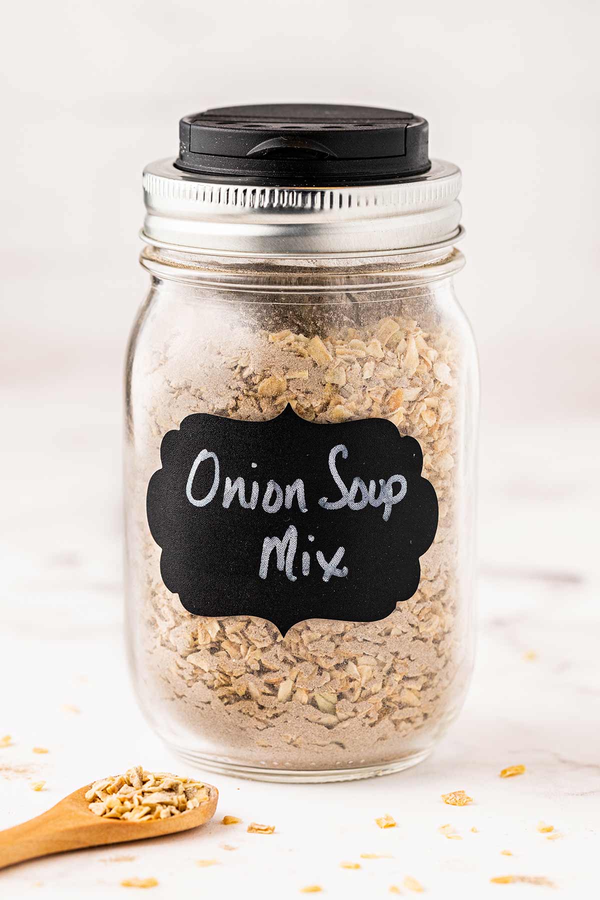 Pint mason jar containing copycat lipton onion soup mix, with chalkboard label on front and wooden scoop laying on marble background.