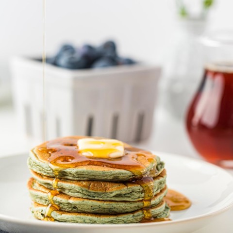 White plate with stack of 4 leftover oatmeal blender pancakes topped with butter and syrup, basket of blueberries and jar of syrup in background.