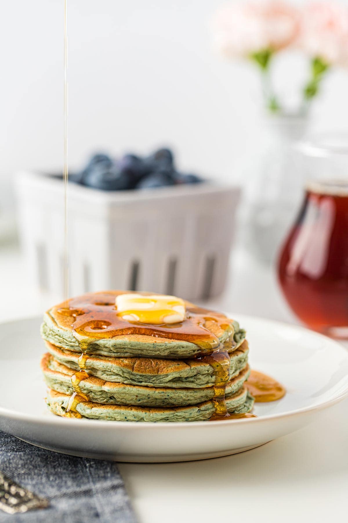 White plate with stack of 4 leftover oatmeal blender pancakes topped with butter and syrup, basket of blueberries and jar of syrup in background.